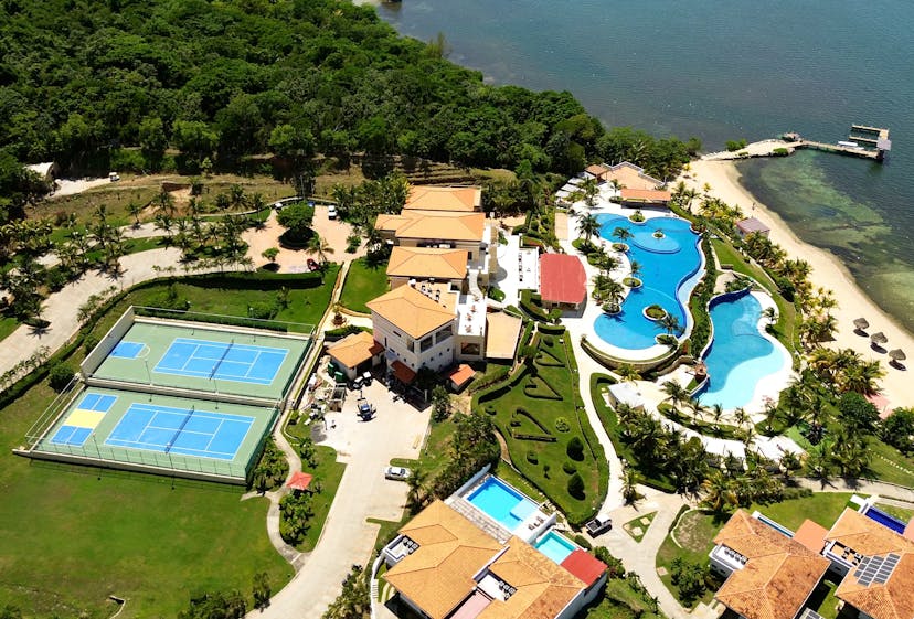 Vista aérea de un complejo turístico de lujo con dos pistas de tenis azules, una gran piscina de forma libre y varios edificios con tejados de terracota rodeados de exuberante vegetación. El complejo está situado junto a una masa de agua con una playa de arena y un muelle que se adentra en el agua.