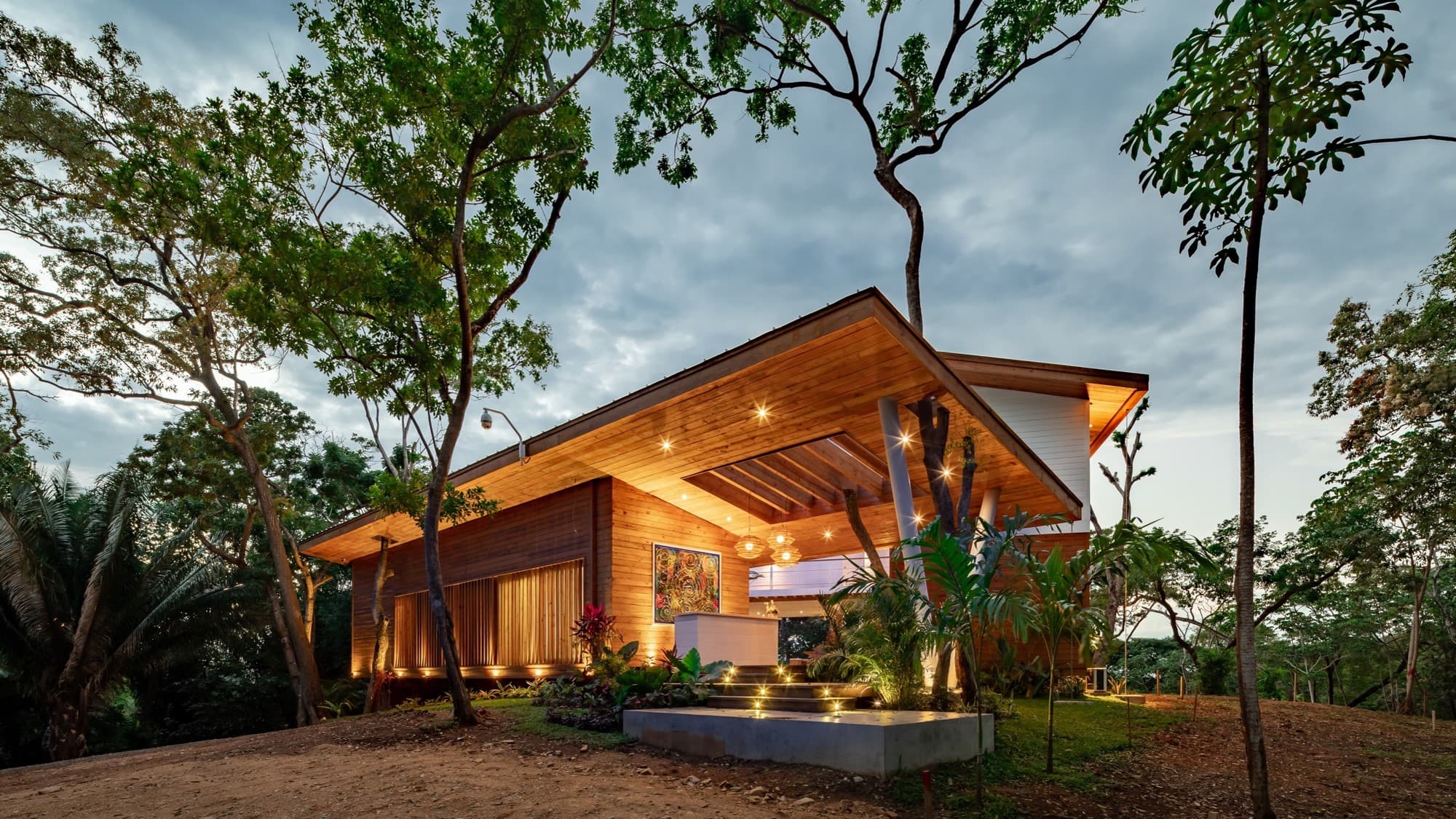 Moderna casa con exterior de madera y amplio tejado en voladizo, bellamente iluminada al atardecer. La casa está rodeada de exuberantes plantas y árboles tropicales, que crean un ambiente sereno y natural.