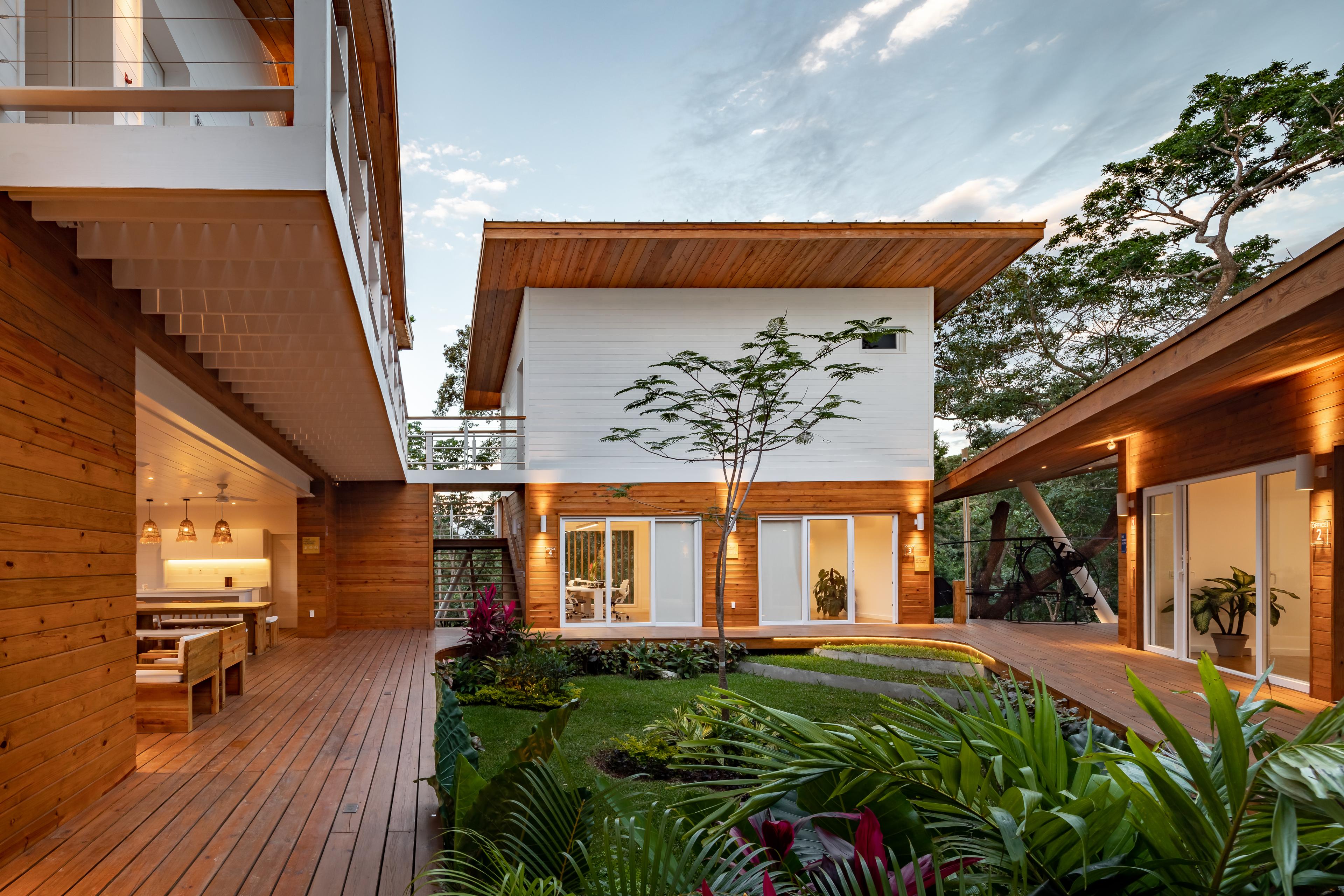 Una casa contemporánea con un exterior de madera que presenta un patio abierto con frondosas plantas y árboles. La casa tiene grandes puertas y ventanas acristaladas, que permiten que la luz natural ilumine los espacios interiores, y la cubierta de madera conecta varias partes de la casa, creando una experiencia de vida interior-exterior sin fisuras.