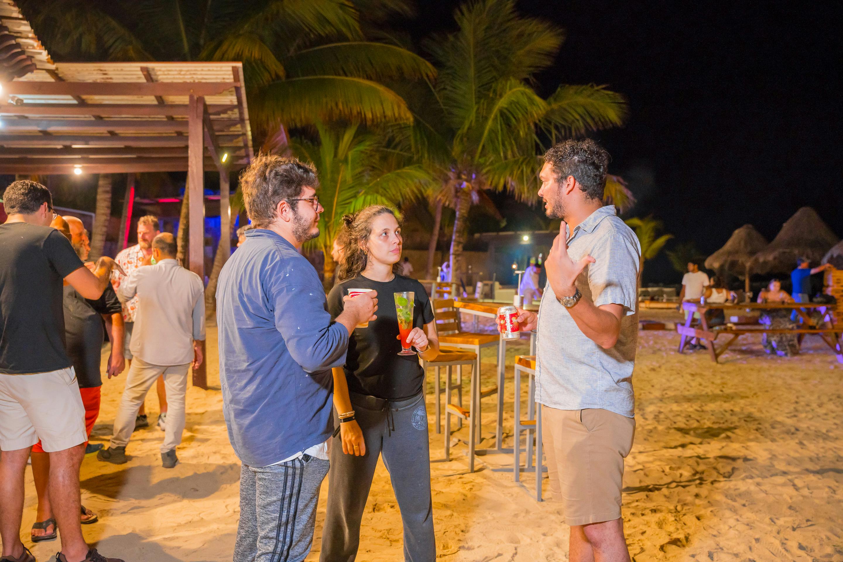 Grupo de personas en una reunión social hablando en una playa con bebidas en la mano.