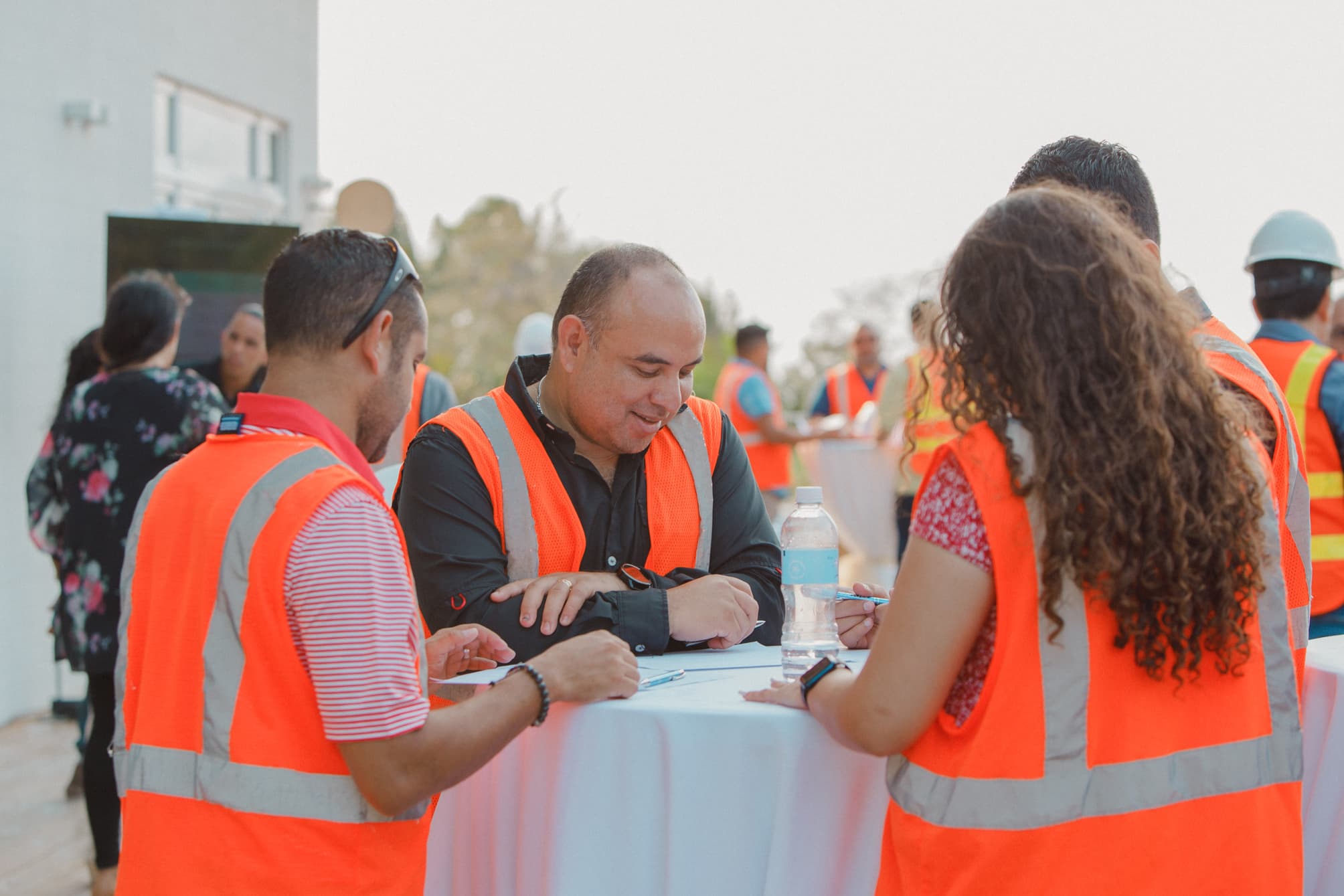 Un grupo de personas con chalecos de seguridad naranjas, reunidas en torno a una mesa y dedicadas a debatir o planificar. El escenario parece estar al aire libre, con algunos participantes sosteniendo documentos y otros interactuando en el fondo, lo que sugiere un entorno de trabajo colaborativo.