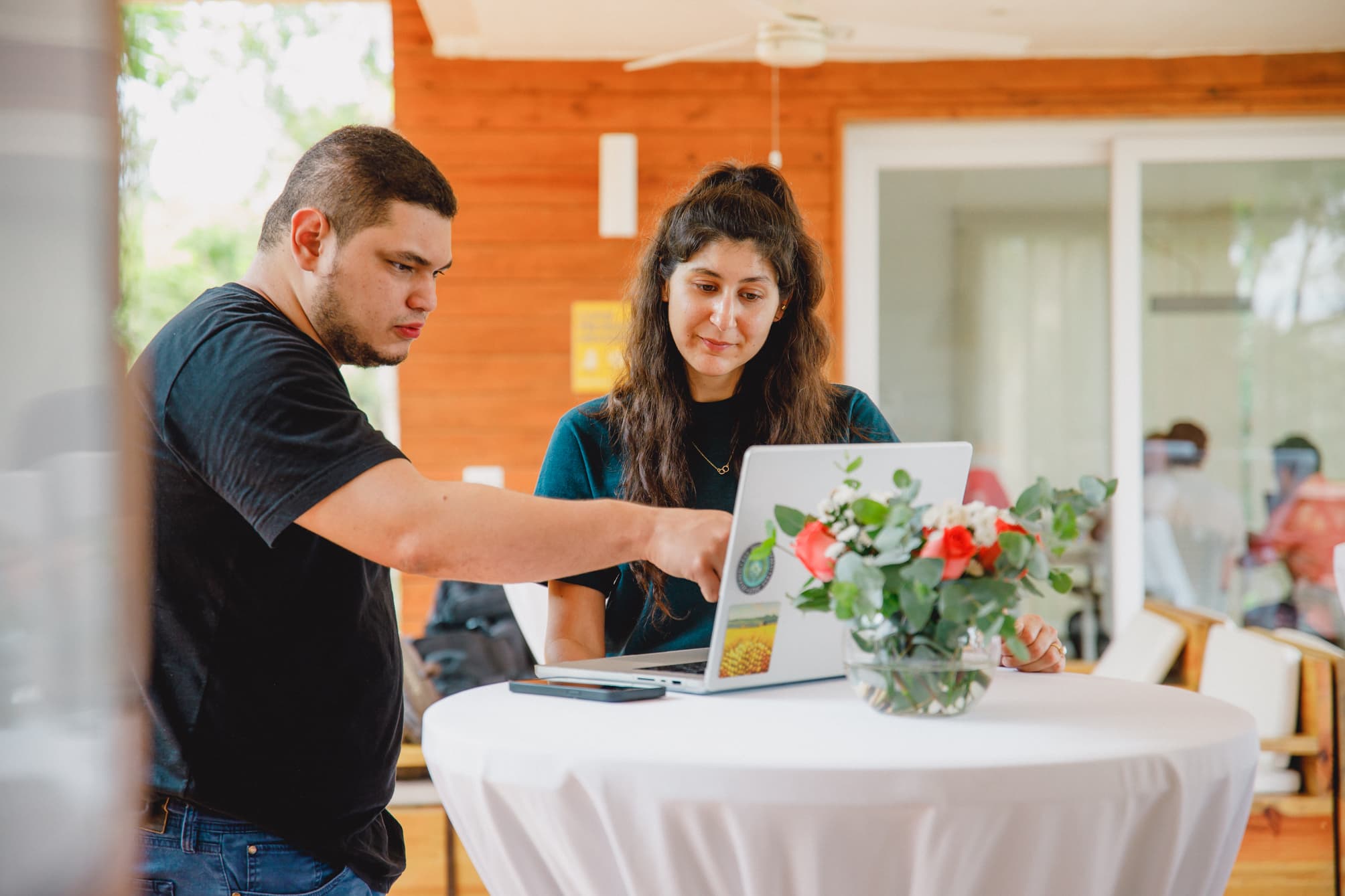 Dos personas de pie ante una mesa alta, con un ordenador portátil abierto frente a ellas. Una de ellas apunta a la pantalla mientras la otra mira atentamente. Un jarrón con flores decora la mesa, y el fondo presenta un interior de madera, creando un ambiente informal y de colaboración.