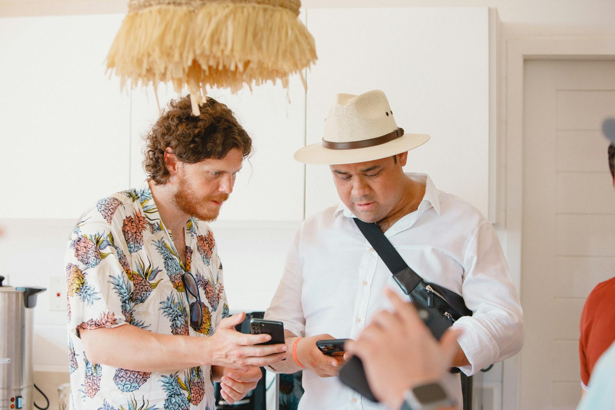 Dos hombres de pie en un interior, mirando atentamente sus teléfonos inteligentes, uno con un sombrero de paja y un bolso cruzado, y el otro con una camisa de flores. Parecen estar discutiendo o compartiendo algo en sus teléfonos, creando una atmósfera de compromiso informal.