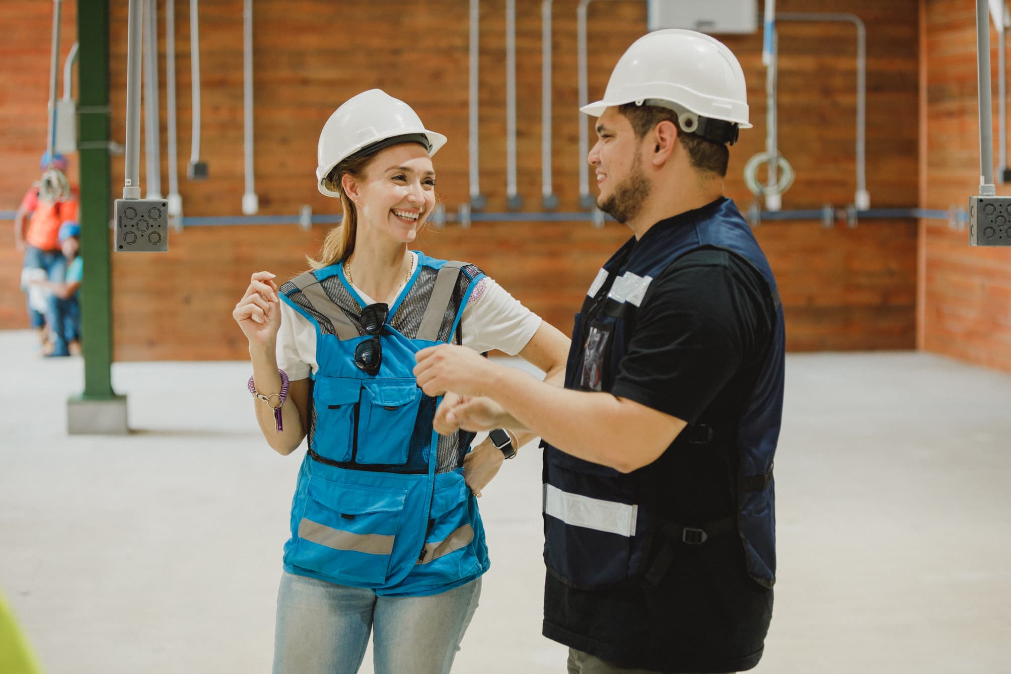 Dos trabajadores de la construcción con cascos y chalecos de seguridad mantienen una conversación en el interior de un edificio en construcción. Sonríen y parecen estar discutiendo algo relacionado con su trabajo, lo que crea un ambiente positivo y de colaboración.