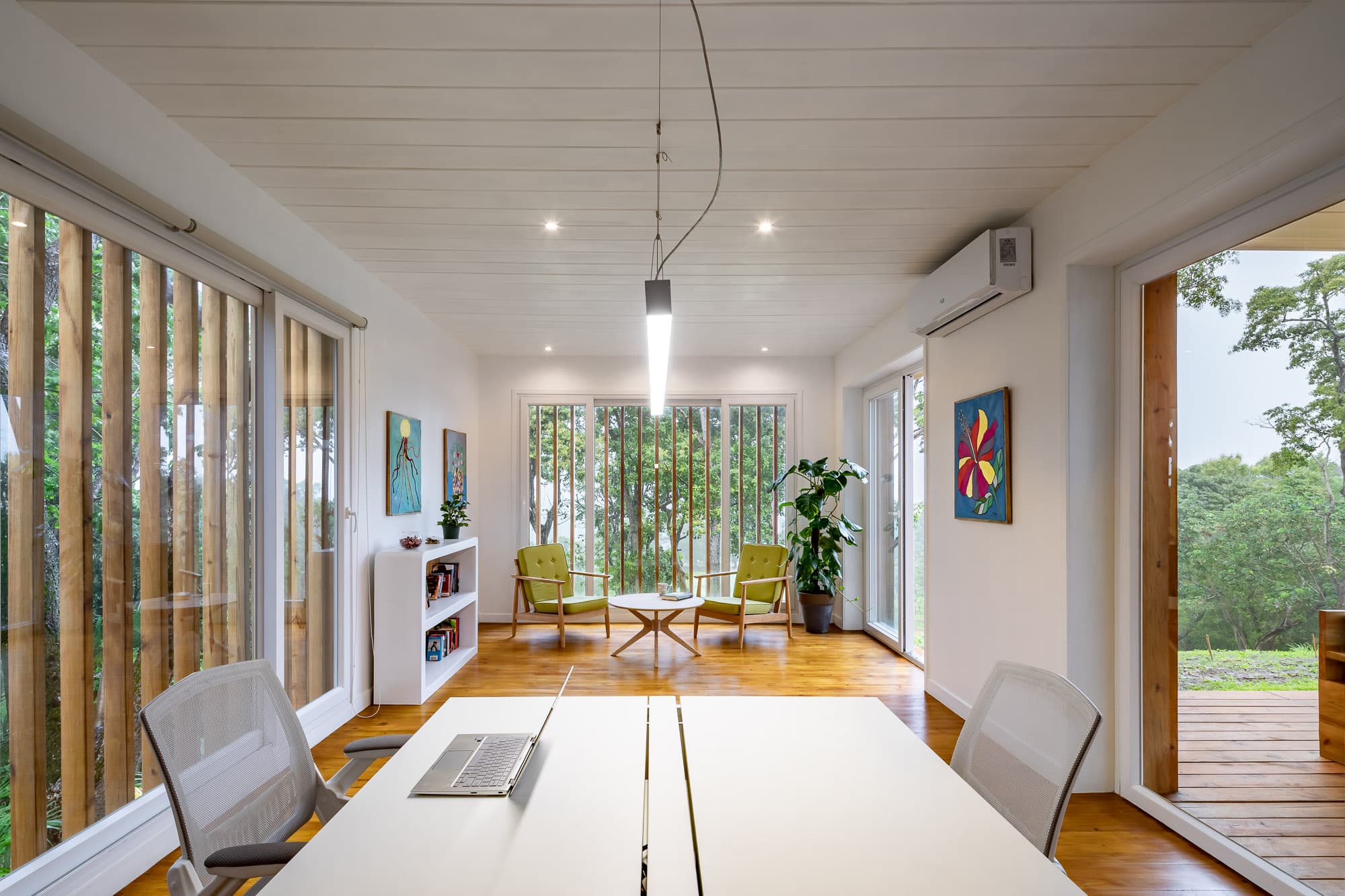 Despacho luminoso y moderno con una gran mesa con un portátil, paredes blancas y suelo de madera. La sala incluye sillas verdes alrededor de una mesa de centro cerca de una gran ventana con persianas verticales, y coloridas obras de arte en las paredes, creando un ambiente acogedor y elegante.