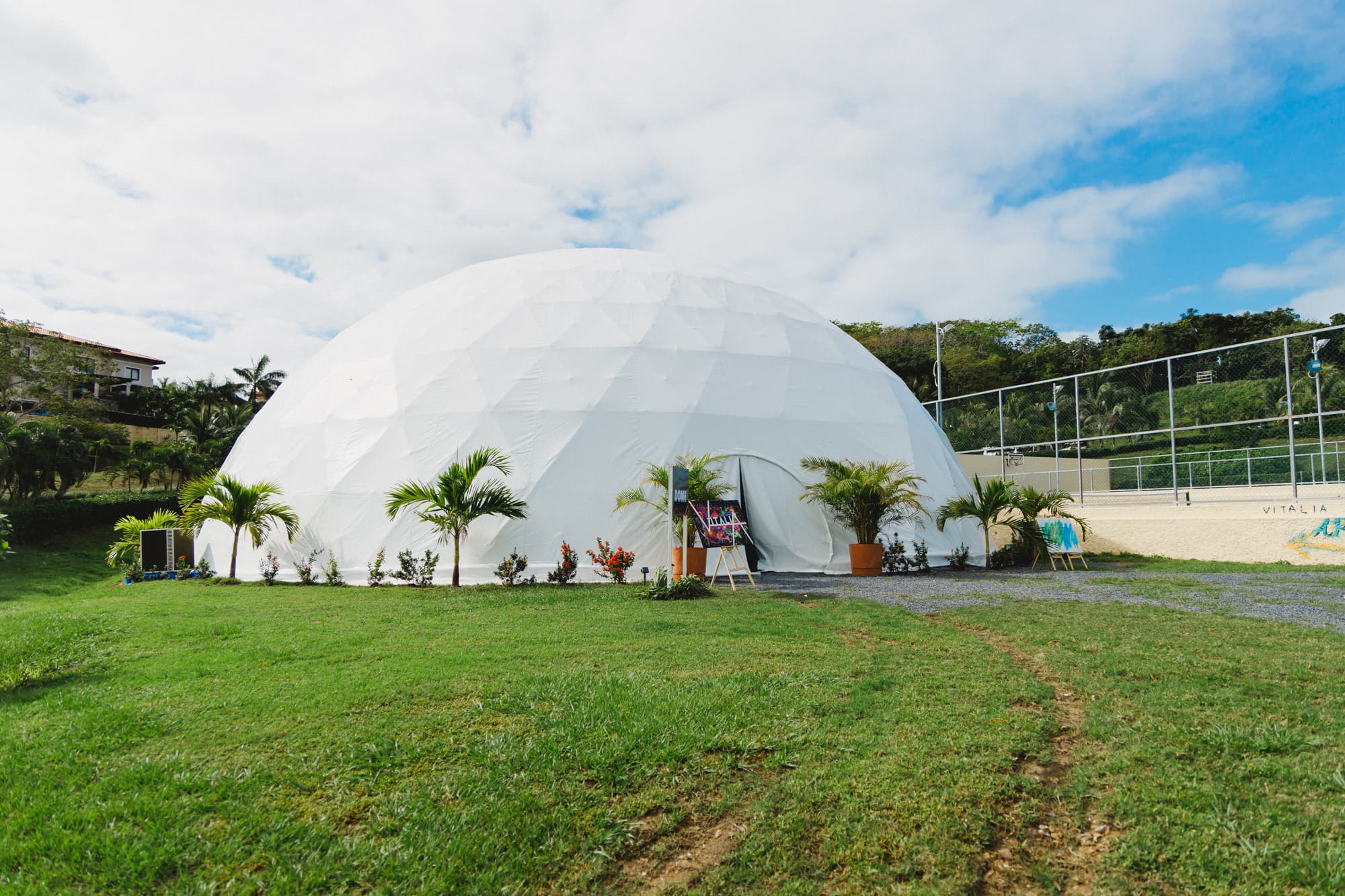 Una gran cúpula geodésica blanca instalada en una zona de césped, con plantas tropicales y pequeños árboles alrededor de la entrada. Es probable que la cúpula se utilice para eventos o reuniones, y el cielo está parcialmente nublado, lo que crea un ambiente luminoso y aireado.