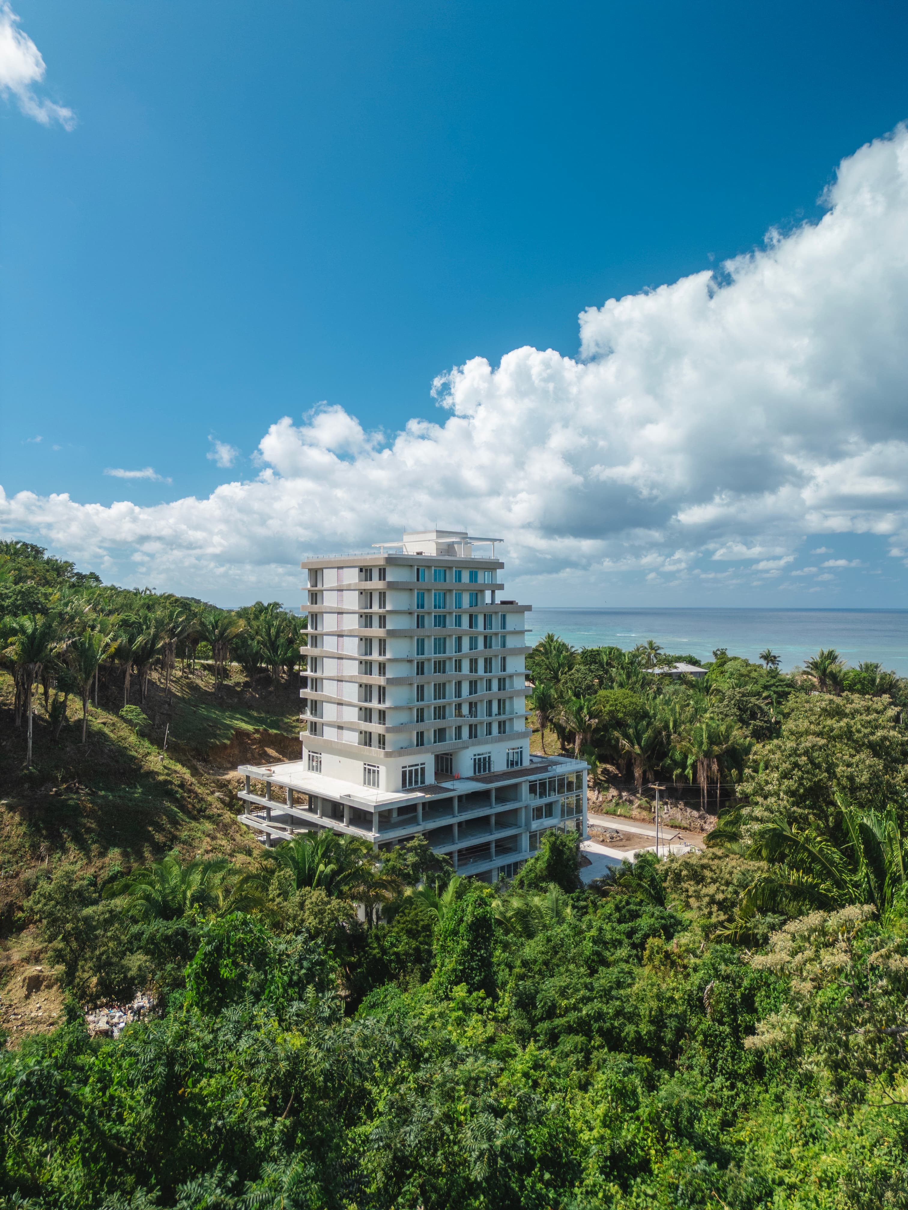 Un edificio alto y moderno en construcción rodeado de vegetación densa y palmeras, con el océano visible al fondo. La estructura destaca sobre el cielo azul con nubes dispersas, lo que indica una ubicación costera con una mezcla de belleza natural e infraestructuras en desarrollo.