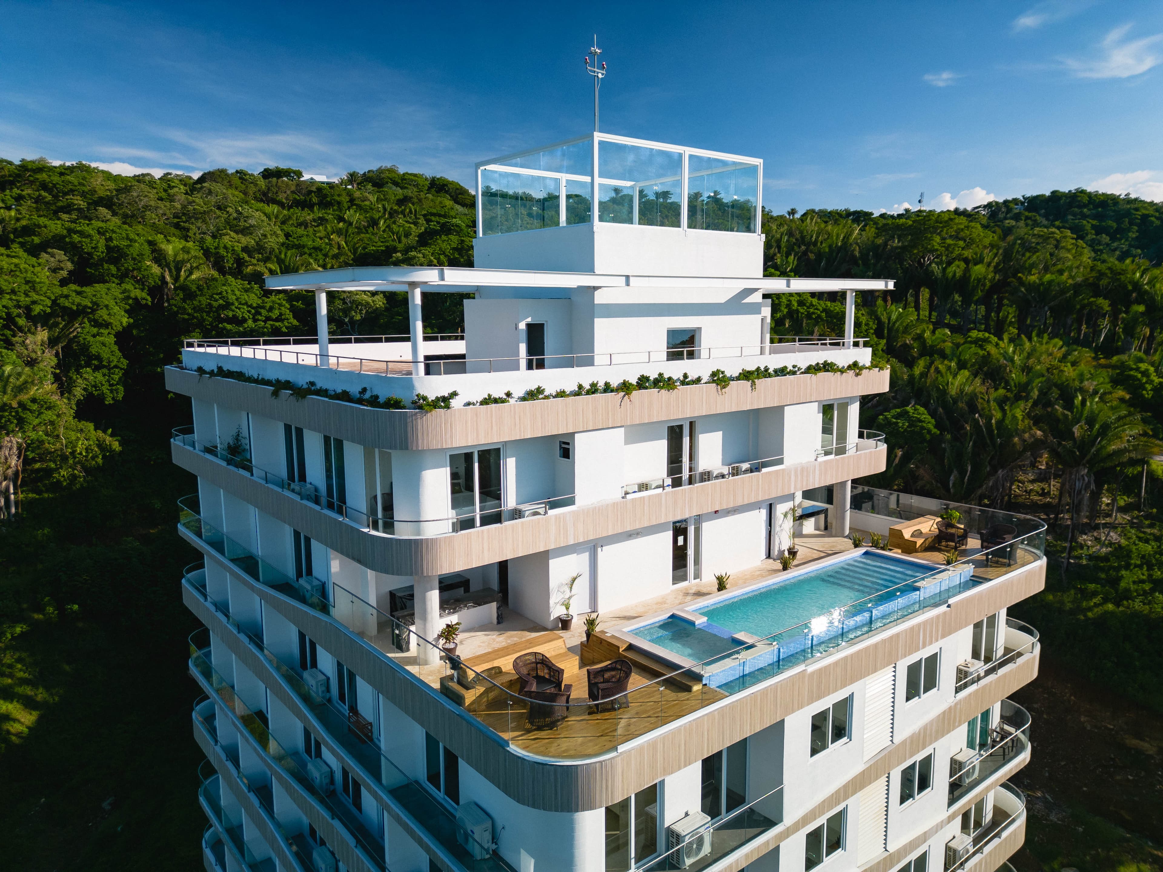 Un moderno edificio de apartamentos o condominios de lujo de varios pisos con paredes exteriores blancas y grandes balcones. En la última planta hay un espacio acristalado. En uno de los balcones inferiores se ve una larga piscina infinita, rodeada de tarima de madera y muebles de exterior. El edificio tiene como telón de fondo un frondoso bosque tropical.