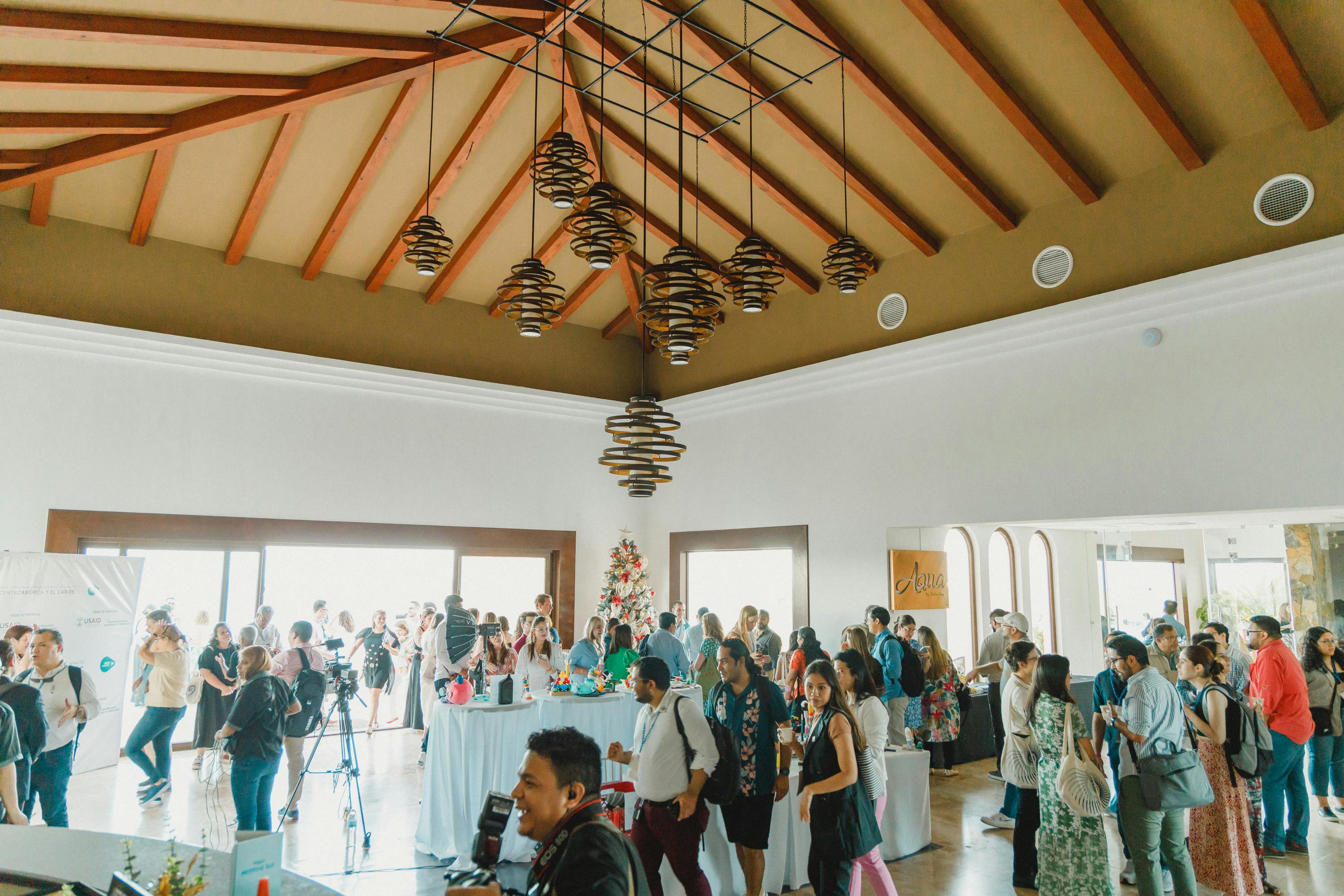 Un grupo numeroso de personas se reúne en una sala espaciosa y bien iluminada, con techos altos con vigas de madera y modernas lámparas colgantes. La multitud parece mezclarse en torno a mesas con bebidas y comida, y algunas personas utilizan cámaras y otros equipos de grabación.