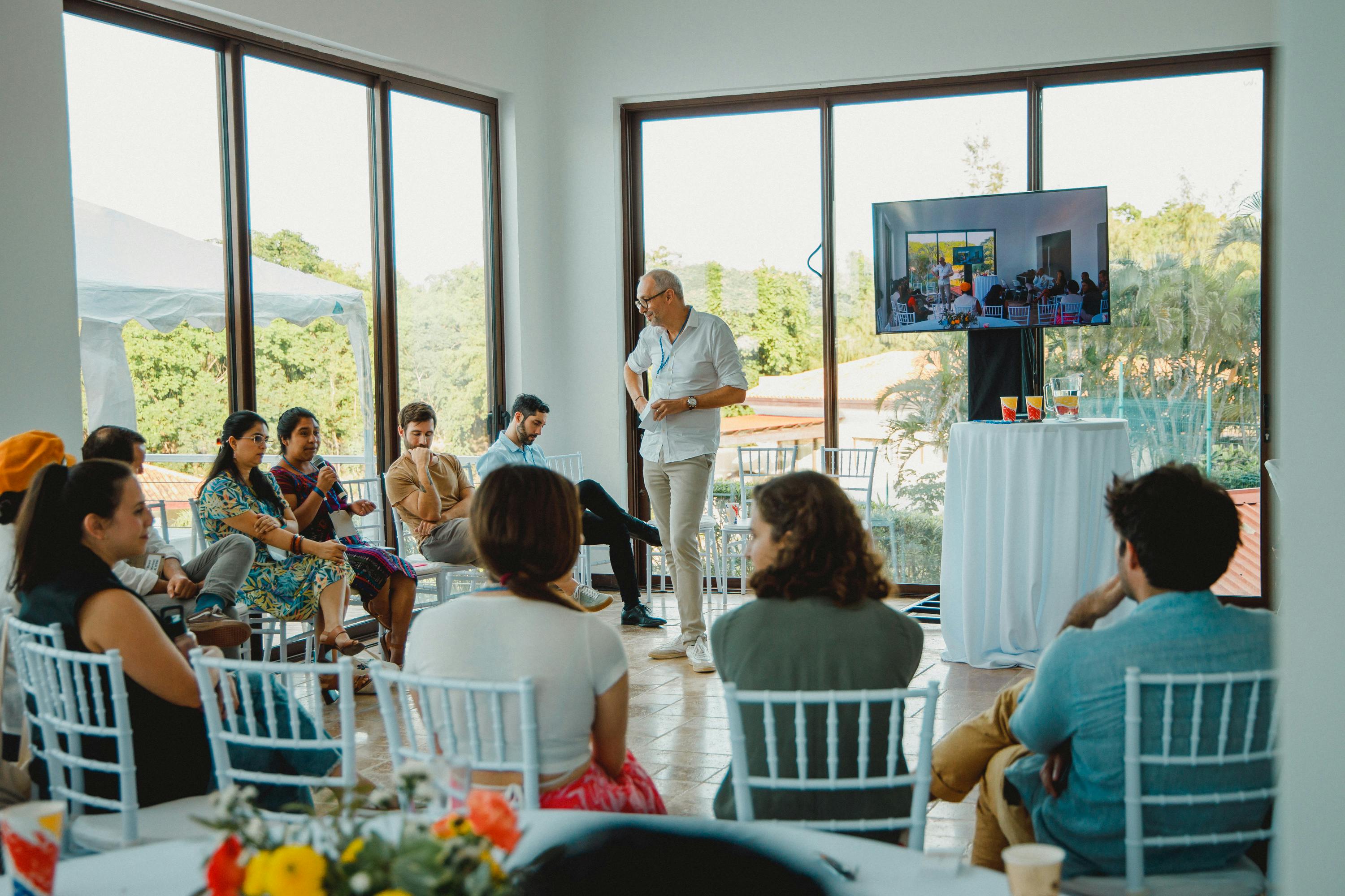 Un grupo de personas sentadas en círculo participan en un debate dirigido por un orador de pie en una sala luminosa con grandes ventanales. Una pantalla de televisión muestra contenidos relacionados y la sala ofrece vistas a la vegetación del exterior, lo que crea un ambiente relajado y atractivo para la reunión.