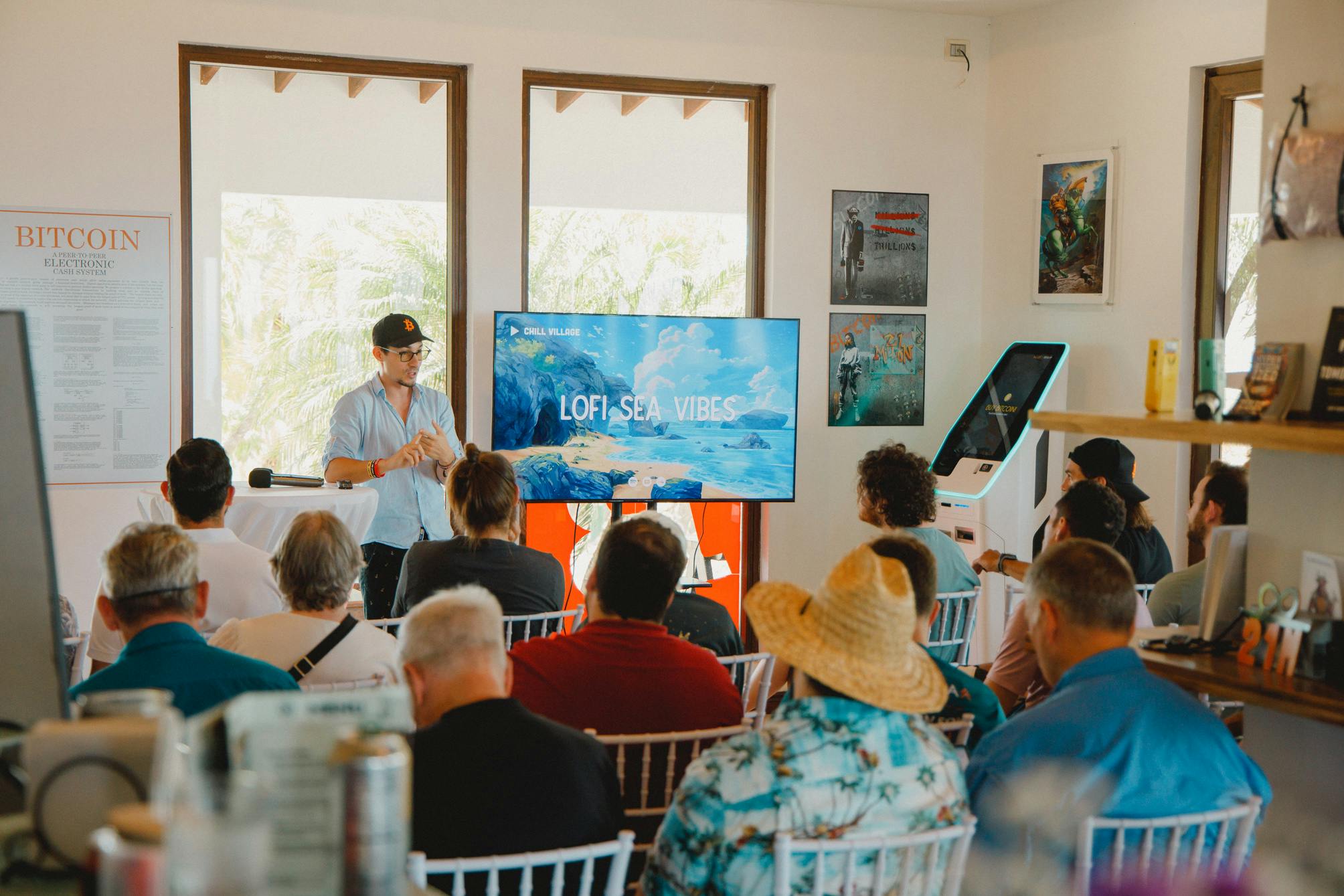 Un grupo de personas sentadas y escuchando a un orador en una sala, con una gran pantalla que muestra LOFI SEA VIBES. Hay carteles en las paredes y grandes ventanales que proporcionan luz natural. El ambiente parece informal y concentrado, con el público involucrado en la presentación.