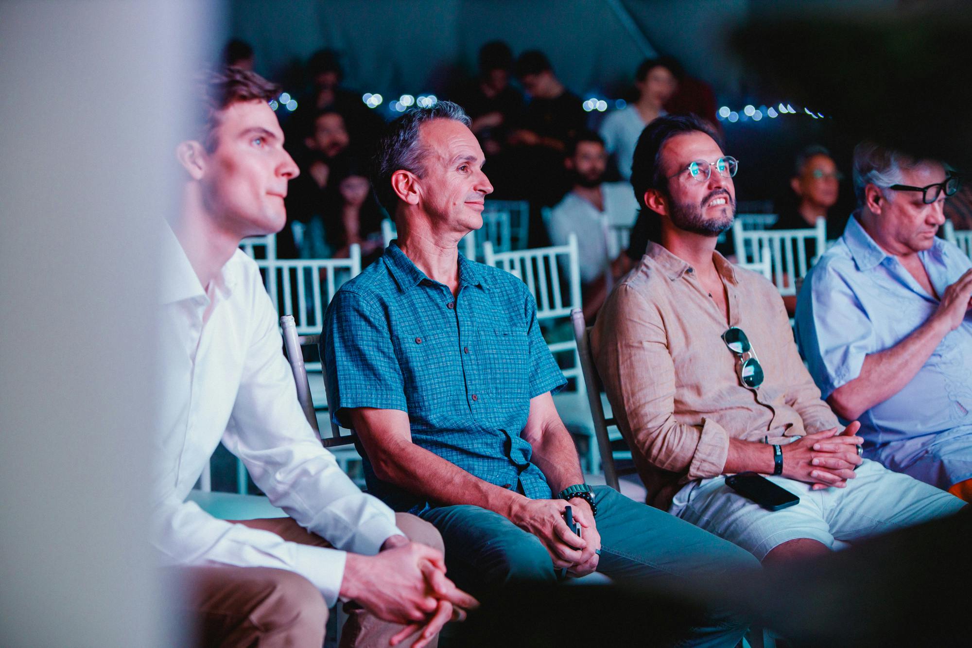 Tres hombres sentados en fila, observando atentamente un acontecimiento, con una multitud al fondo. El escenario parece ser un interior con un enfoque en los asistentes de la primera fila.