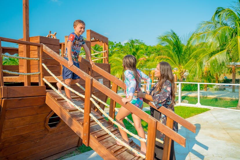 Un niño de pie sobre el puente de madera de una estructura de juego, sonríe a una niña y a una mujer que le animan. La zona de juegos está ambientada al aire libre, con palmeras y un cielo azul despejado de fondo.