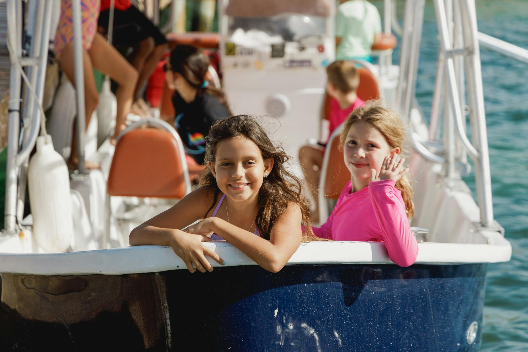 Dos chicas sonríen y disfrutan de un paseo en barco; una de ellas saluda a la cámara. Al fondo se ven otros pasajeros, lo que crea un ambiente animado y alegre en el agua.