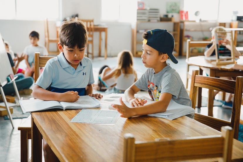 Dos niños sentados a una mesa de madera en un aula, enzarzados en una discusión sobre libros y papeles abiertos. Al fondo se ve a otros niños trabajando en un entorno de aprendizaje colaborativo y relajado.