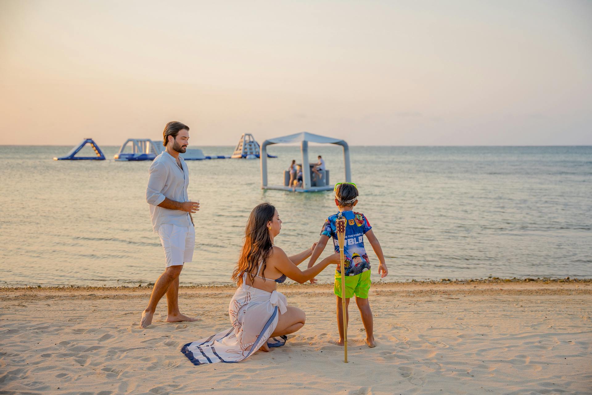 Un padre y una madre con un niño en una playa al atardecer, con estructuras acuáticas hinchables flotando al fondo. El mar en calma y el cielo en tonos pastel crean un ambiente tranquilo y relajado, perfecto para una tarde junto al agua.