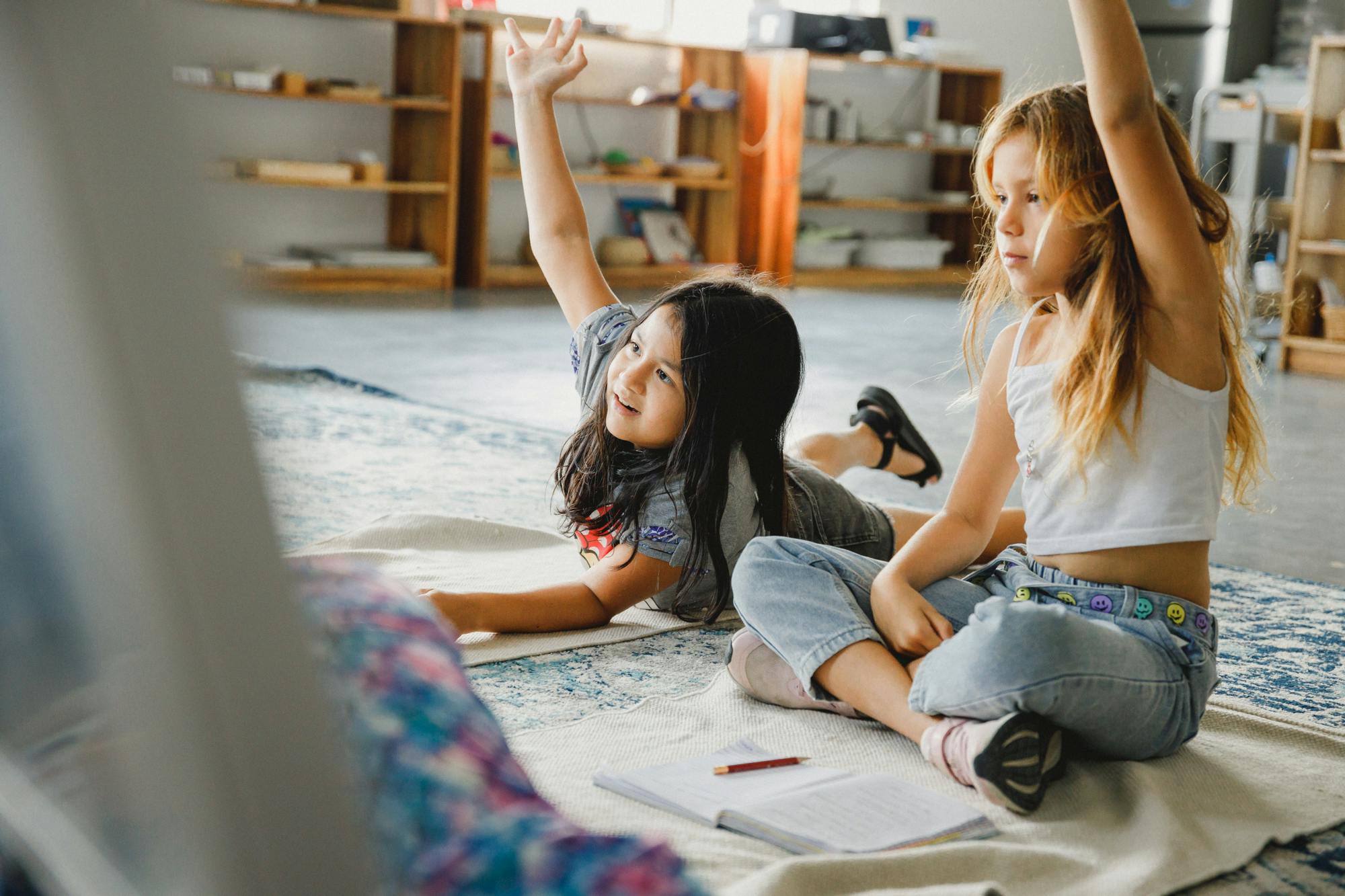 Dos niñas sentadas en el suelo de una clase, levantando las manos y participando en una lección. El ambiente es relajado, con libros y materiales esparcidos y estanterías llenas de recursos educativos al fondo.