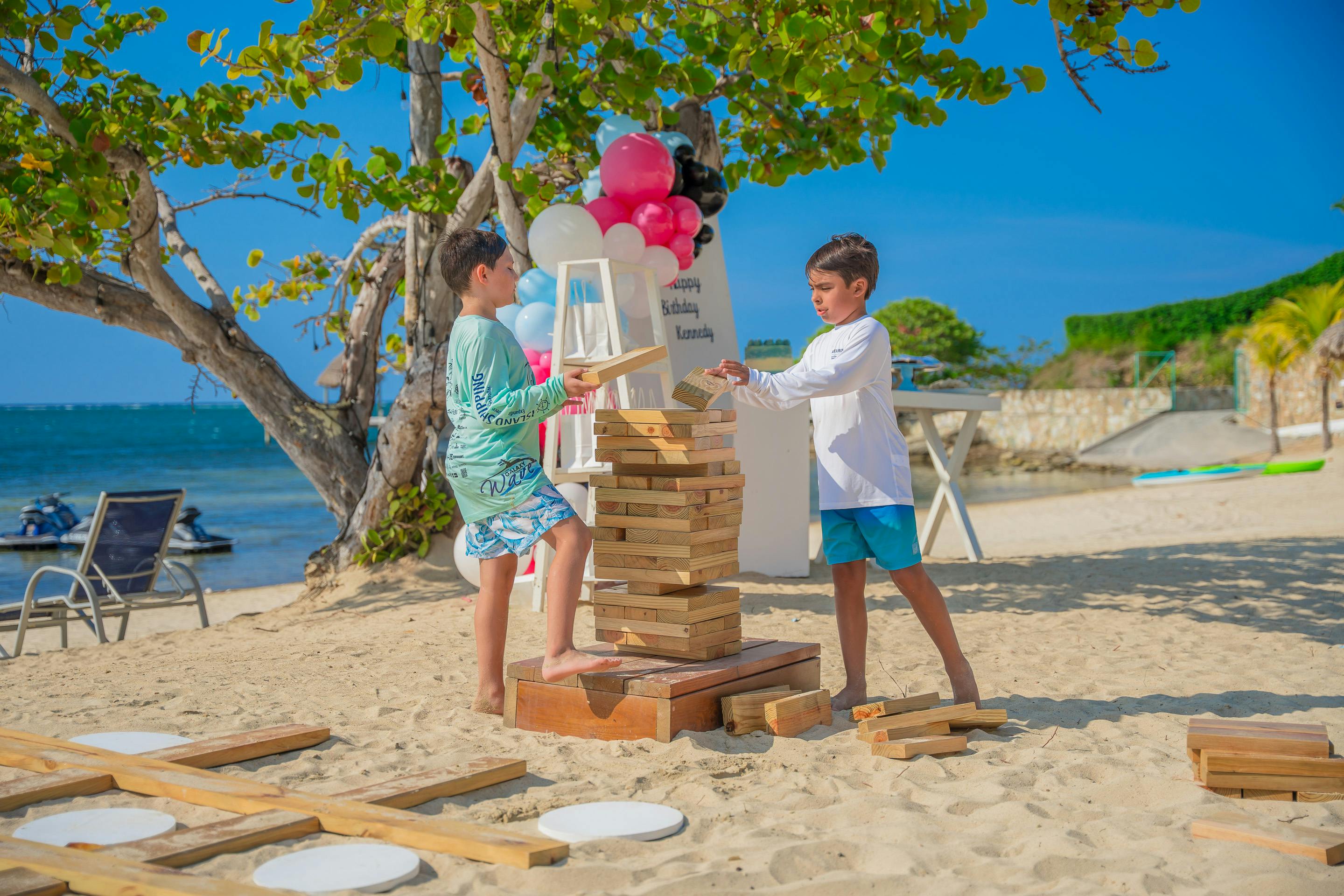 Dos niños juegan una partida de Jenga gigante en una playa de arena, con árboles y globos de fondo. La escena es luminosa y soleada, con un montaje festivo con decoraciones coloridas y una hermosa vista del océano de fondo.