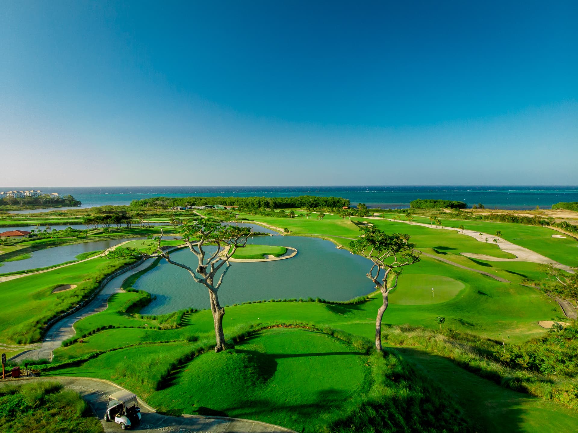 Vista aérea de un campo de golf con greens bien cuidados, trampas de arena y varios obstáculos de agua, con el océano visible en la distancia. El campo es exuberante y verde, con un cielo azul despejado que crea un paisaje sereno y pintoresco.