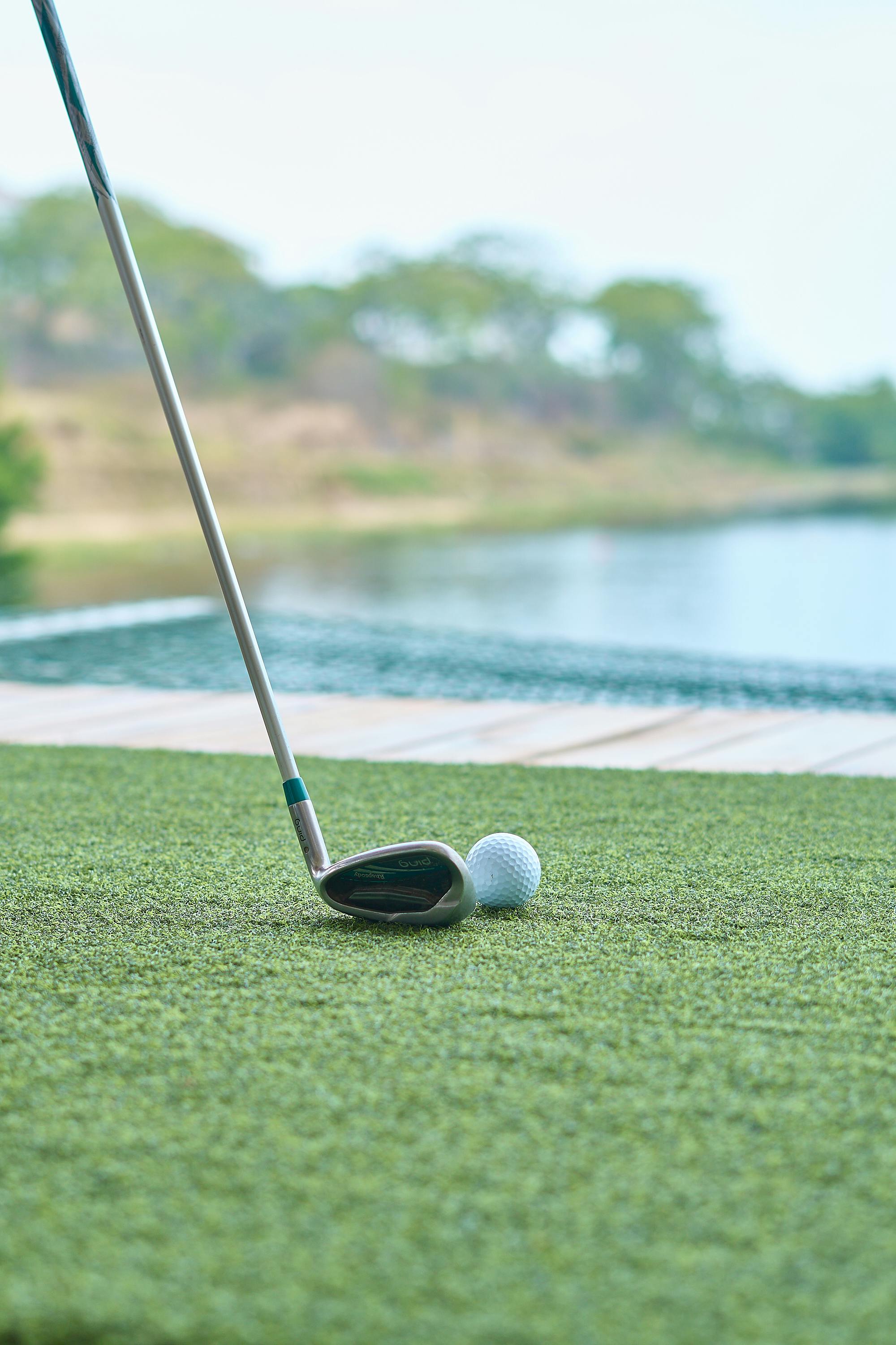 Primer plano de un palo y una pelota de golf sobre césped artificial cerca de un obstáculo de agua en un campo de golf. La calma del agua y la vegetación del fondo crean un entorno tranquilo y concentrado para un partido de golf.