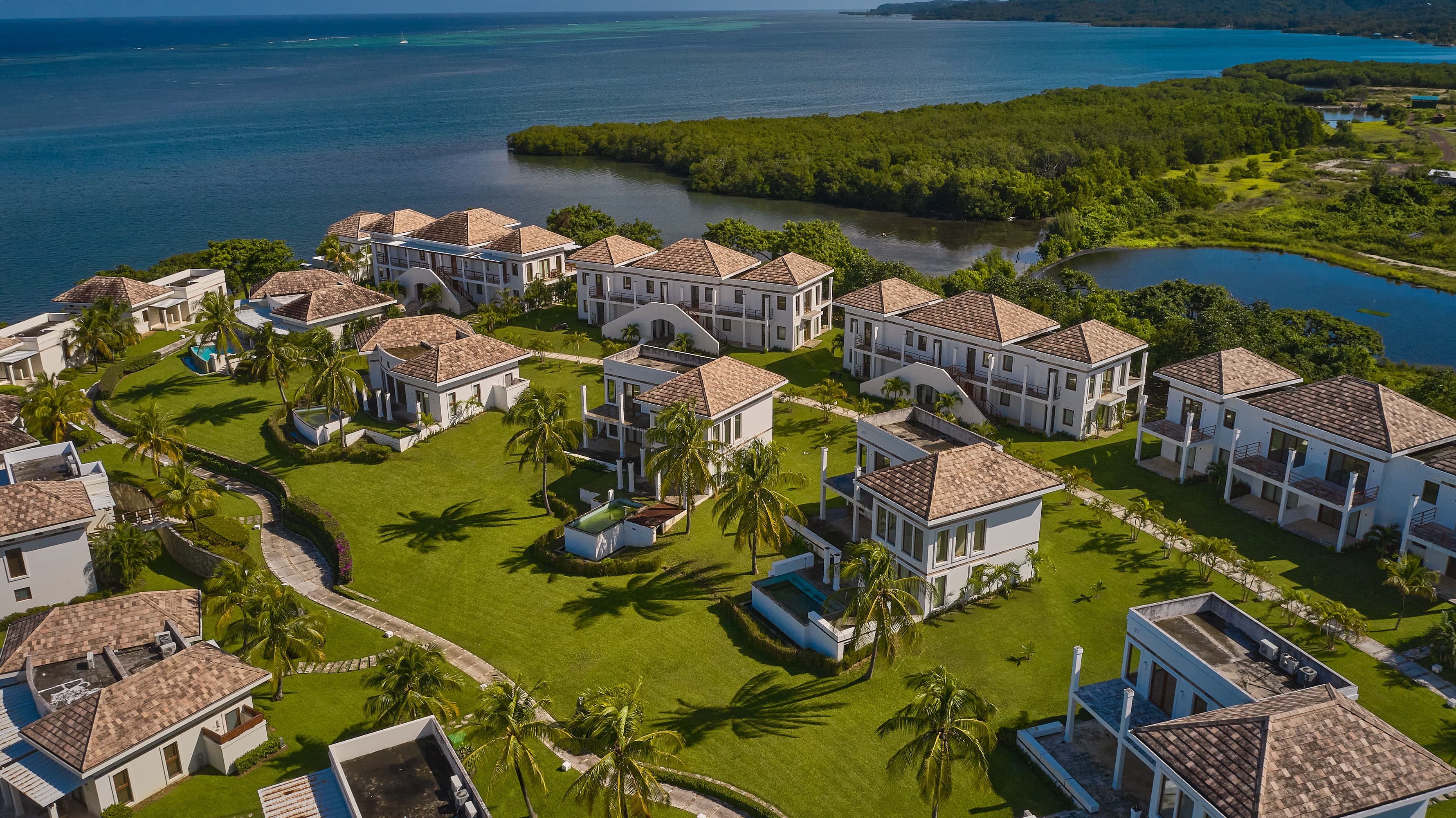 Vista aérea de un complejo turístico frente al mar con varias villas de dos plantas con tejados de tejas, rodeadas de césped bien cuidado y palmeras. Las villas están situadas a lo largo de una pintoresca costa de aguas tranquilas y exuberante vegetación, creando un entorno tranquilo y lujoso.