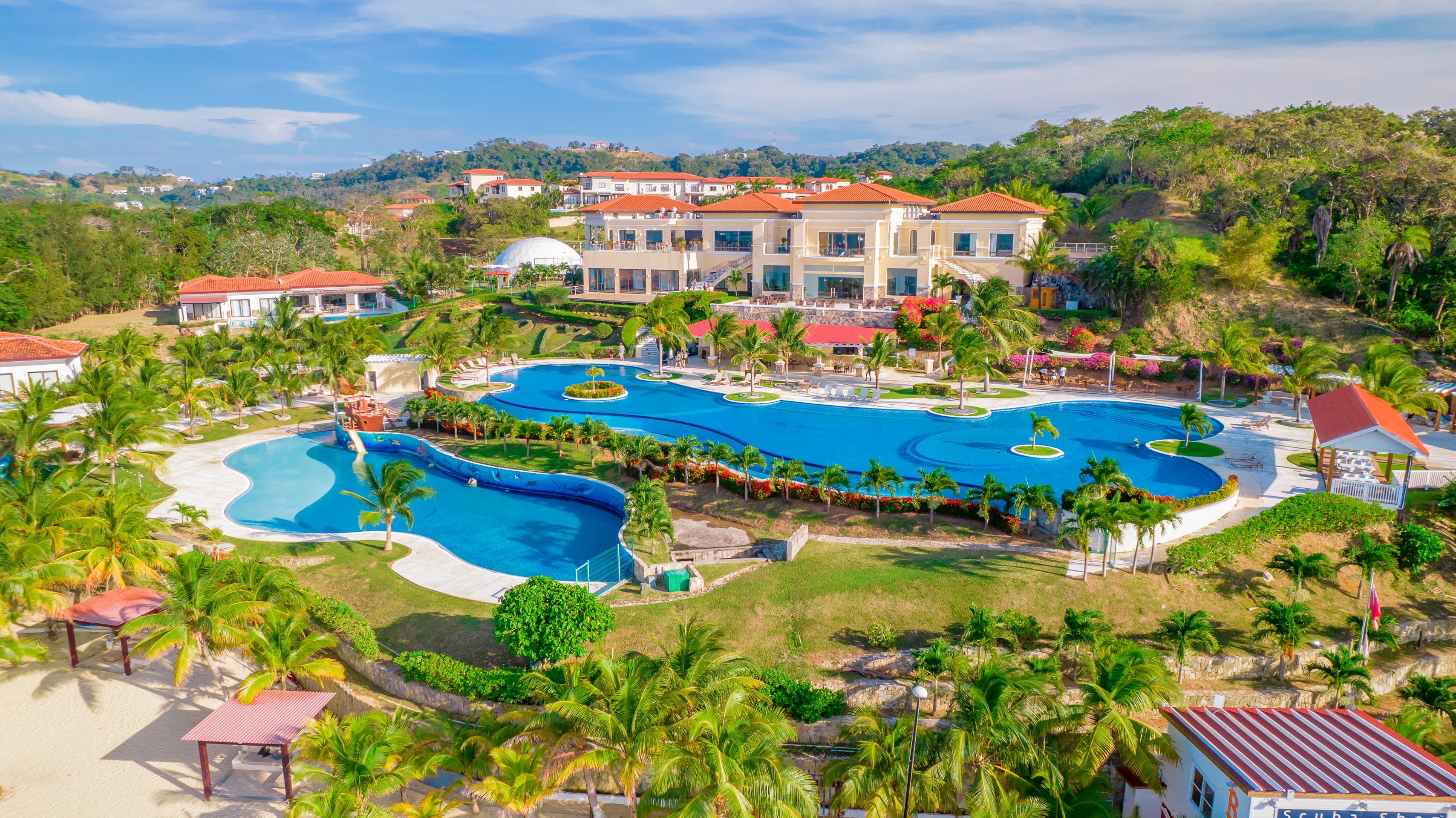 Vista aérea de un complejo turístico con una gran piscina serpenteante rodeada de exuberante vegetación y palmeras. Los edificios del complejo tienen tejados de terracota y dan a la zona de la piscina, con colinas y otras villas visibles al fondo.