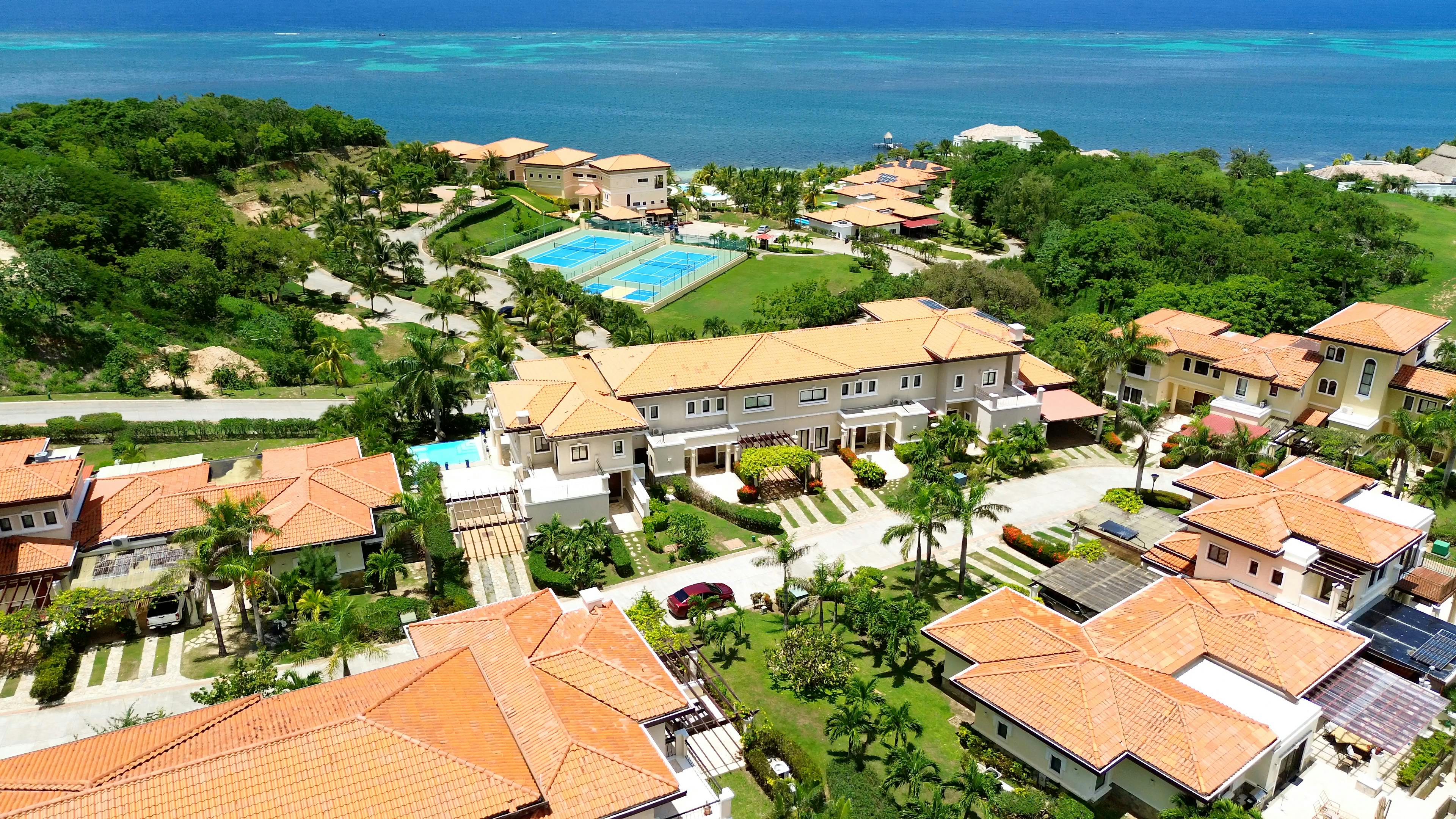 Vista aérea de un complejo turístico costero con varios edificios con tejados de terracota, pistas de tenis y un exuberante paisaje verde. El complejo está situado junto a un océano azul, con la playa y un muelle visibles al fondo, creando un entorno pintoresco y lujoso.