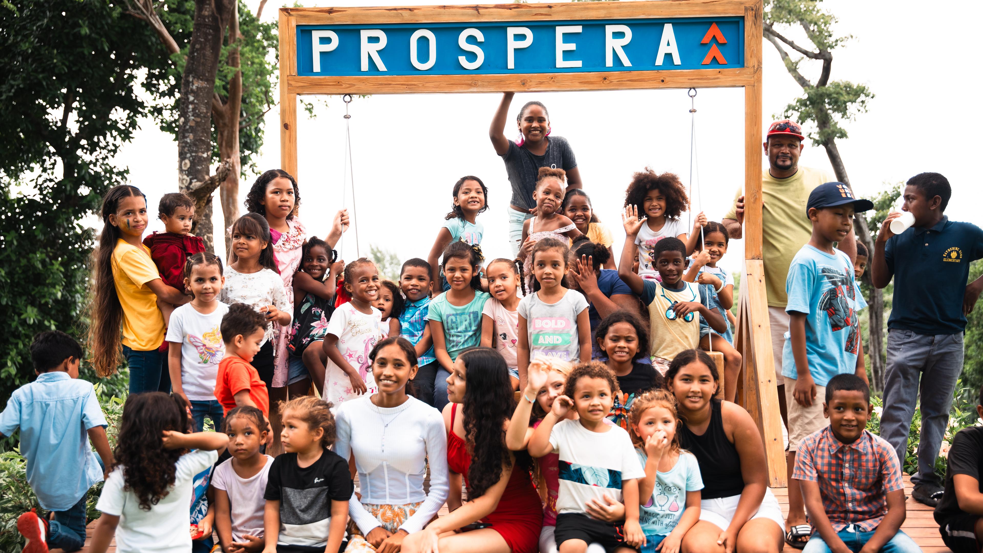 Un grupo numeroso de niños y algunos adultos reunidos bajo un cartel de madera en el que se lee PROSPERA, sonríen y posan para la cámara. Parece que se trata de un acto comunitario o una celebración, y todos parecen alegres y comprometidos.