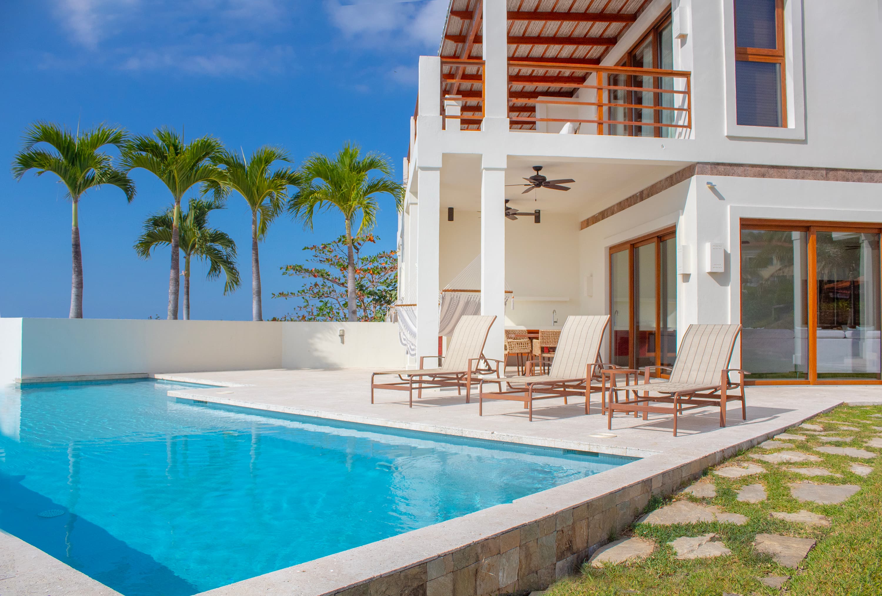 Lujosa villa frente al mar con piscina privada, con un moderno exterior blanco, amplio patio con tumbonas y palmeras contra un cielo azul claro. Una propiedad ideal para alquileres vacacionales en el trópico, con servicios de lujo y una vida interior y exterior.