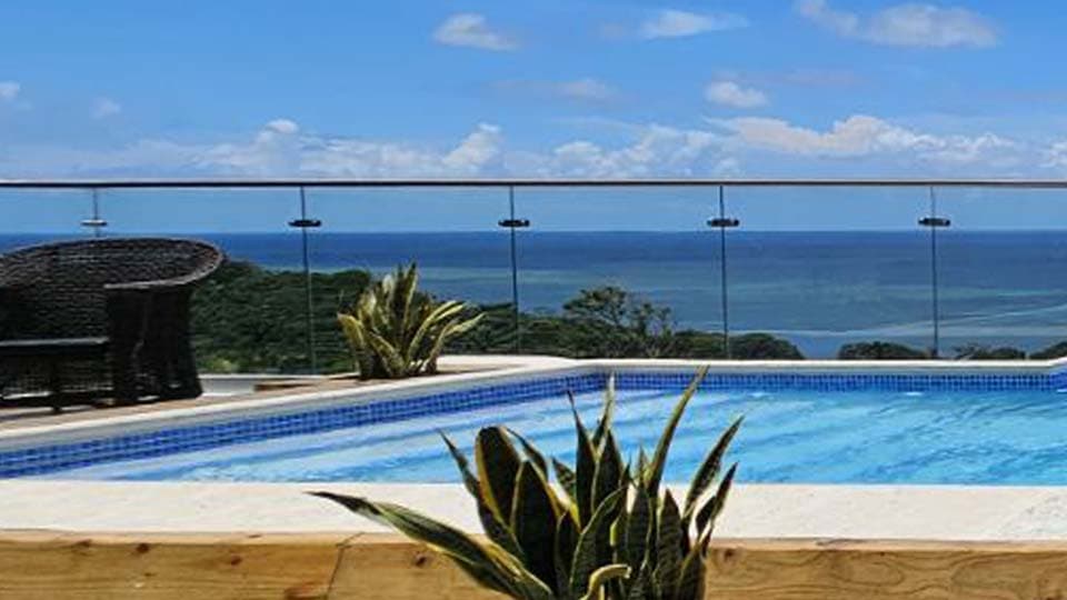 Elegante salón costero con vistas panorámicas al océano a través de grandes puertas correderas de cristal. Cuenta con un amplio sofá seccional gris con almohadas azules y blancas, mesa de centro de madera y suelos de baldosas pulidas. Diseño interior luminoso y aireado perfecto para casas de lujo frente al mar o alquileres vacacionales de alto nivel.