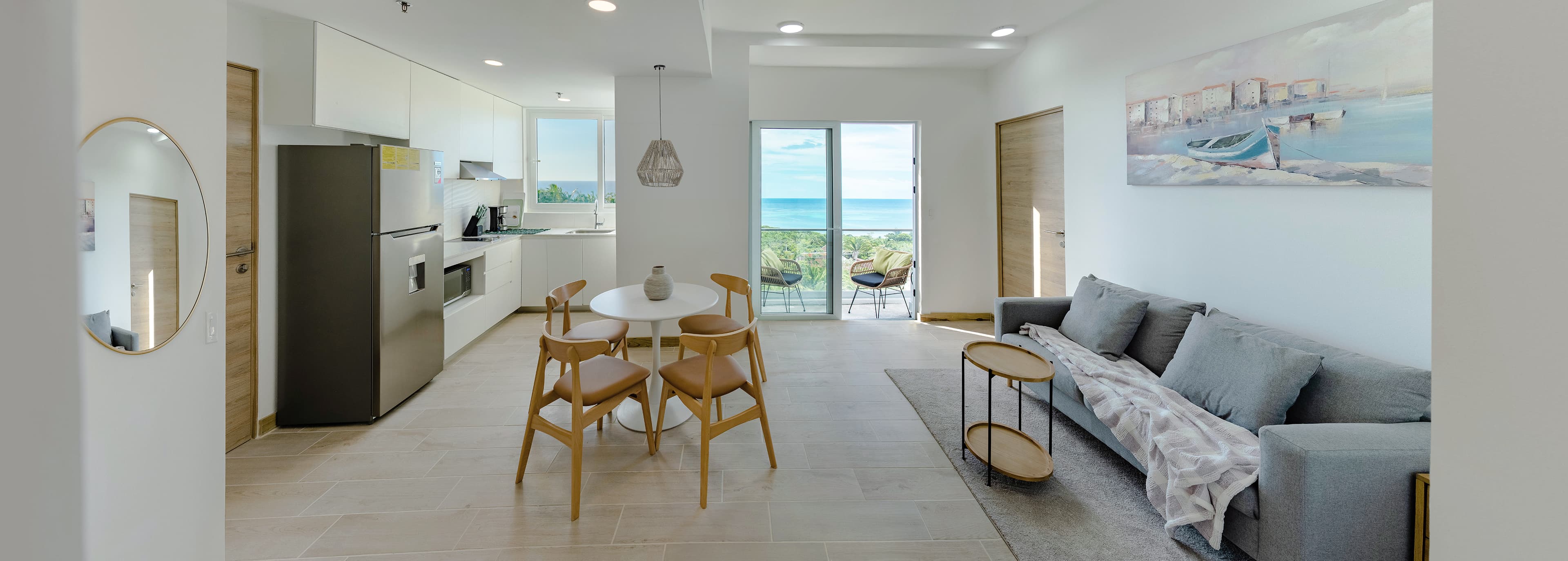 Moderno apartamento frente al mar con sala de estar de planta abierta con cocina blanca totalmente equipada, elegante juego de comedor y cómodo sofá gris. Los grandes ventanales ofrecen impresionantes vistas al océano, mientras que la decoración de temática costera y un balcón privado realzan el ambiente junto al mar. Ideal para alquileres vacacionales de lujo o para una vida costera contemporánea.