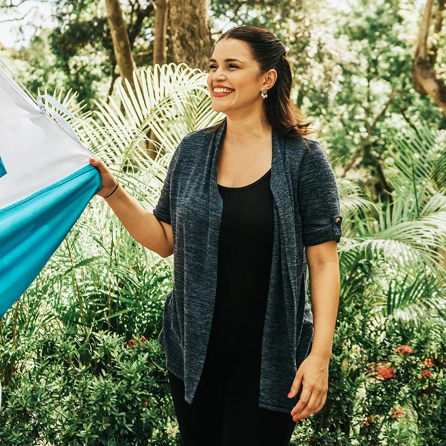 Una mujer sonríe y sostiene una bandera azul y blanca al aire libre, rodeada de frondosa vegetación y árboles. Va vestida con una rebeca azul marino sobre un top negro, creando una escena alegre y natural.