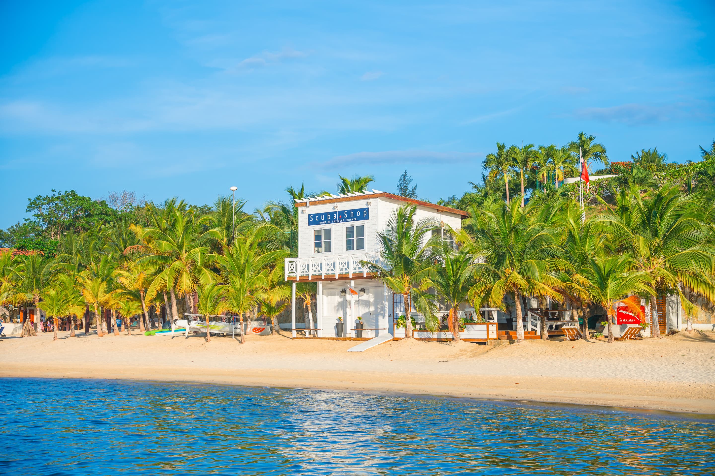 Un edificio blanco de dos plantas con la etiqueta Scuba Shop situado en una playa de arena, rodeado de palmeras. El edificio da a un océano azul y tranquilo, creando una escena pintoresca y acogedora perfecta para los amantes de la playa y el submarinismo.