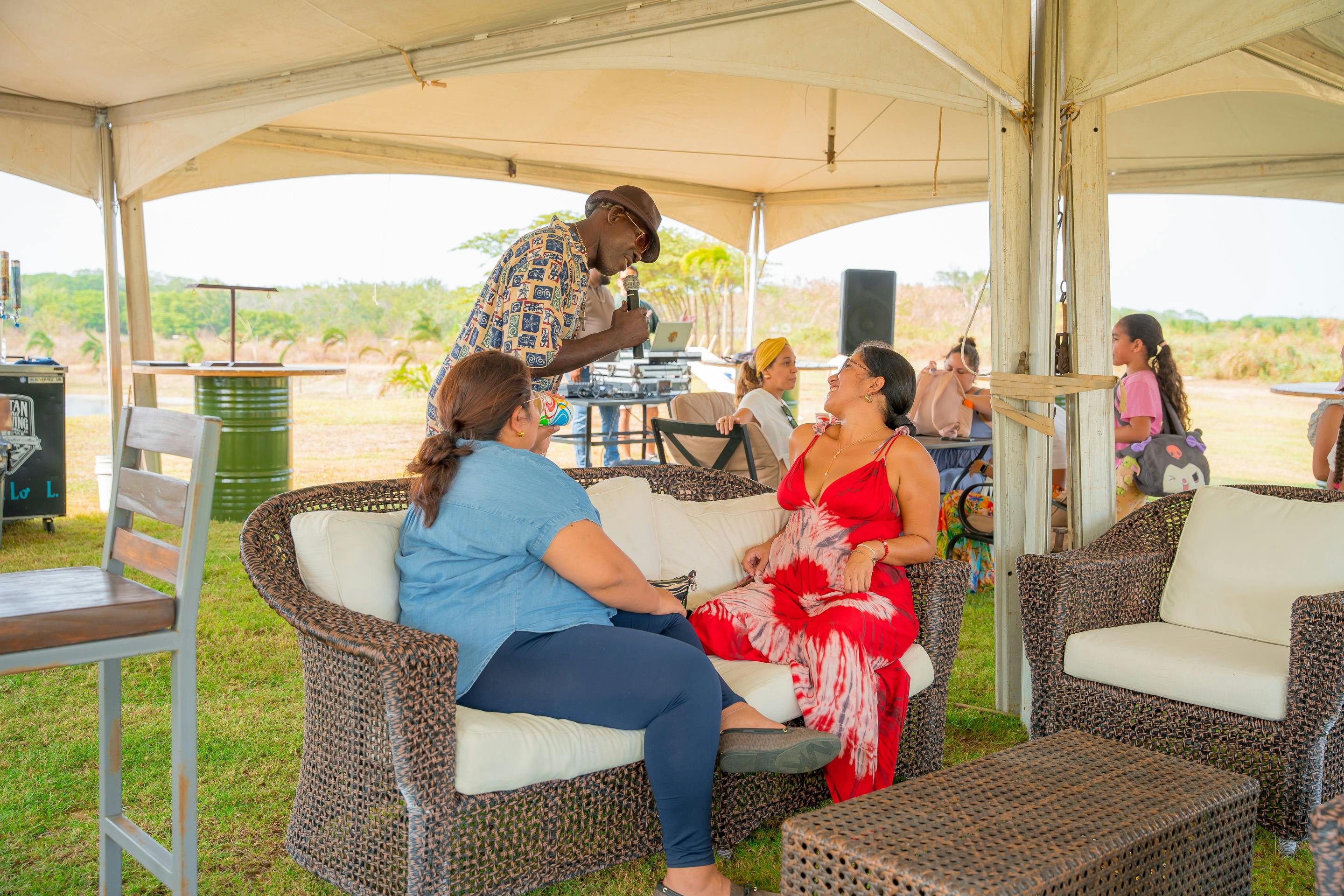 Un grupo de personas sentadas en muebles de mimbre bajo una carpa, entablan una animada conversación. Un hombre habla por un micrófono, creando un ambiente relajado y social en un entorno al aire libre.