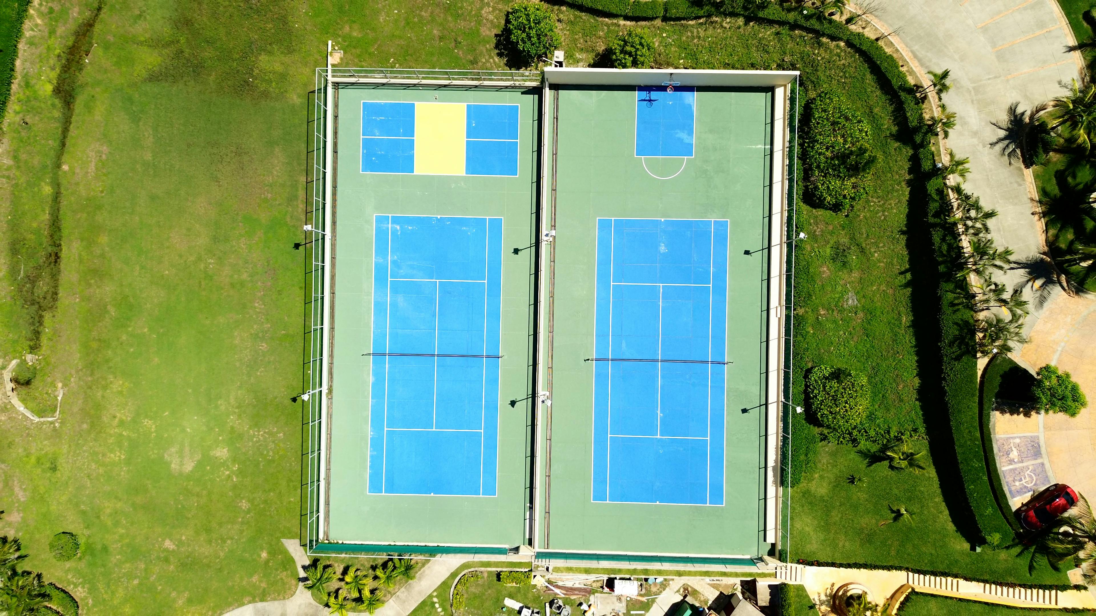 Vista aérea de dos pistas de tenis adyacentes con superficies azules, rodeadas de césped verde y jardinería. En una de las pistas también hay una zona más pequeña de color amarillo que probablemente se utilice para otro deporte o actividad. Las pistas están bien mantenidas y situadas cerca de una zona de aparcamiento y de un coche rojo.